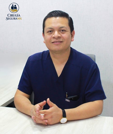 Male healthcare professional in navy scrubs sitting at a desk with hands clasped, smiling at camera