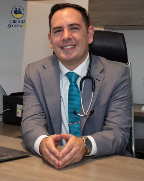 Male doctor in gray suit and teal tie with stethoscope smiling at desk in medical office