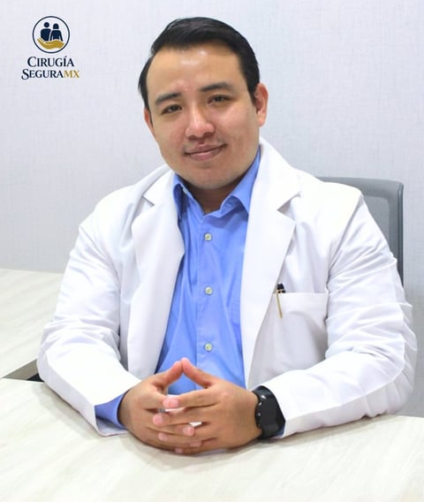 Male doctor in white lab coat and blue shirt sitting at desk with Cirugia Segura MX logo visible in background
